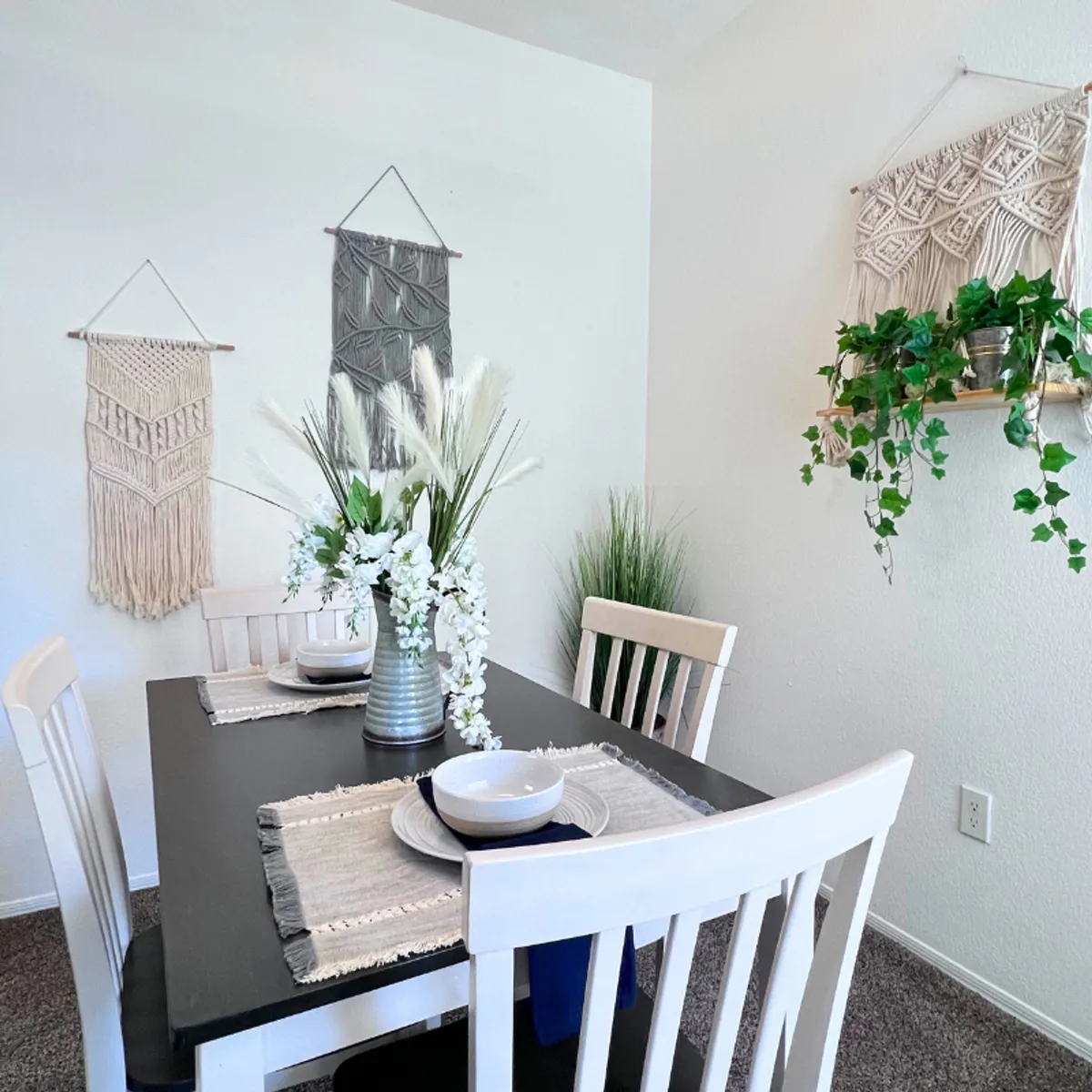 A stylish dining area with a modern table set for a meal, complemented by a vibrant floral arrangement, boho macrame wall hangings, and lush indoor plants. The space radiates warmth and is designed for gathering and relaxing.