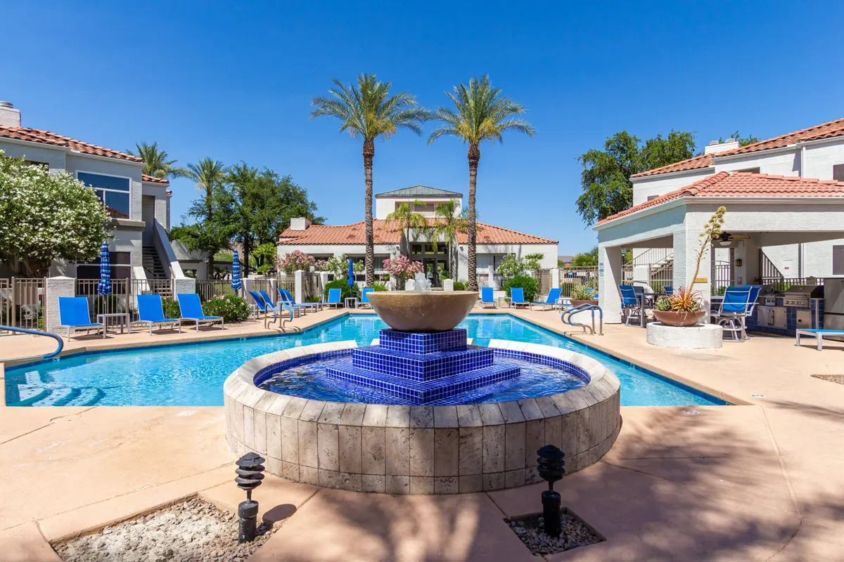 A sparkling swimming pool bordered by a fountain, palm trees, and lounge chairs. The area is inviting for both swimming and relaxing, set in a tranquil and recreational atmosphere.