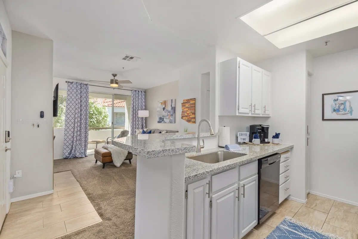 The image reveals a stylish living area with a cozy couch, decorative artwork, and a view of the kitchen counter, evoking a sense of comfort and easy flow between spaces. Soft lighting and curated details create a tranquil, homey environment.