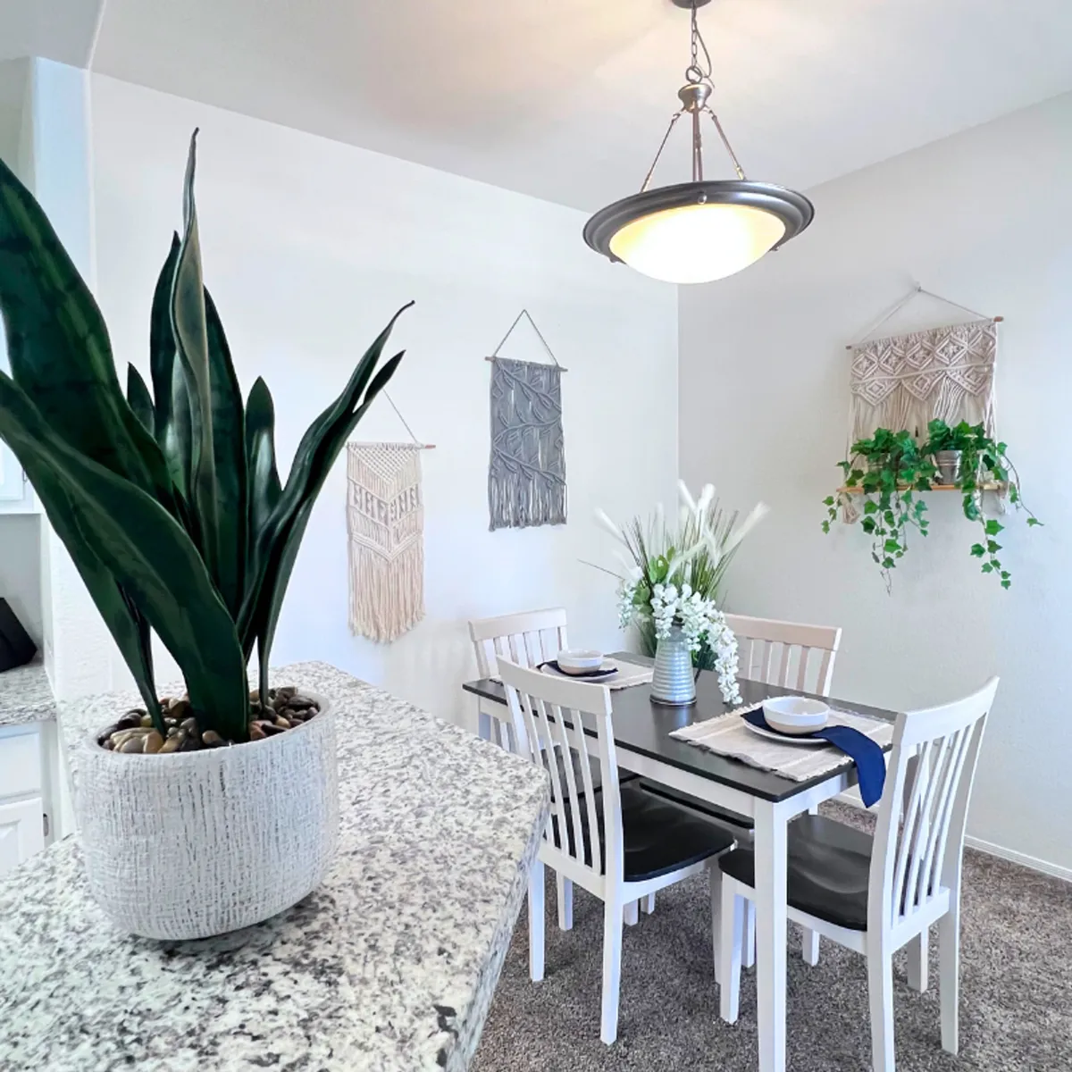 A cozy and welcoming dining nook featuring a dining table, hanging macrame wall art, and a potted plant. The intimate setting is perfect for family meals, entertaining guests, or working remotely in comfort.