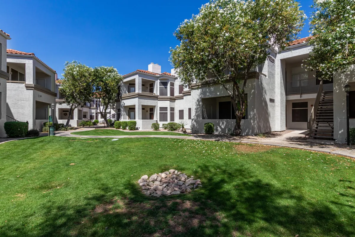 Featuring a lush green lawn, decorative stone accents, shade-giving trees, and glimpses of the building’s architecture, this image showcases a quiet, well-maintained courtyard ideal for peaceful strolls or moments of reflection.